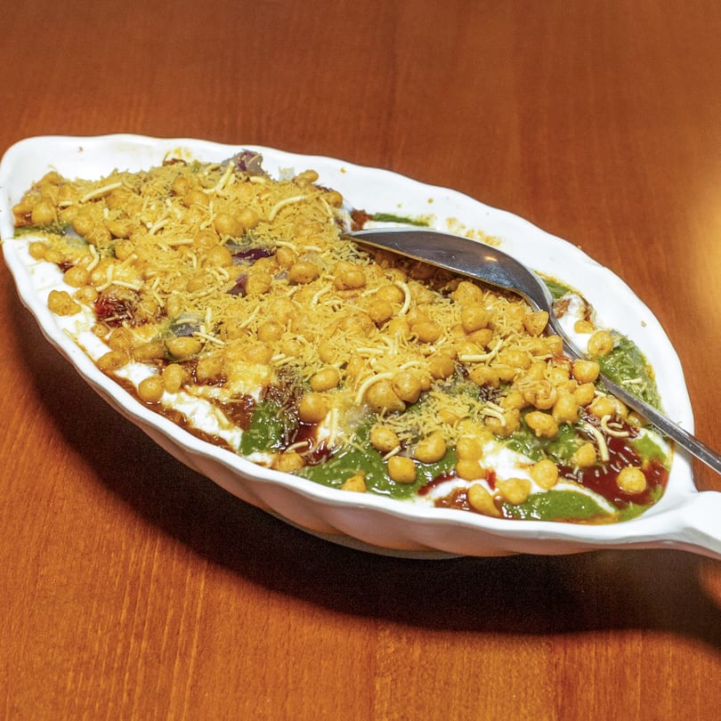 A white platter of Indian chaat with yogurt, green and red chutneys, and crispy toppings.