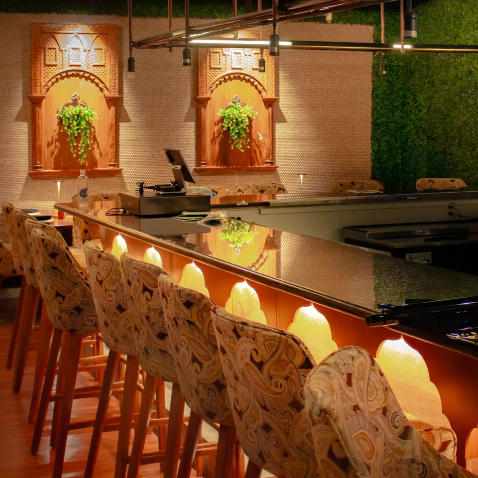 Warmly lit restaurant bar with patterned upholstered stools, ornate wooden wall panels, hanging plants, and reflective countertops.