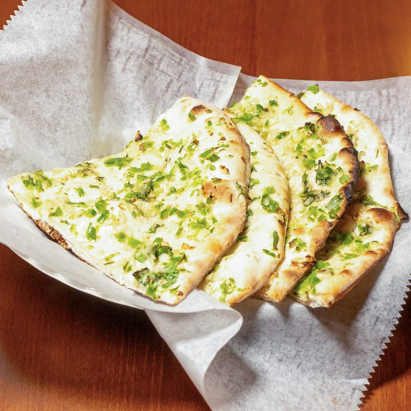 Four pieces of garlic naan bread, topped with green herbs, served in a white paper-lined basket.