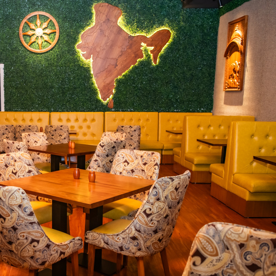 Warmly lit restaurant dining area with mustard-yellow booths, paisley chairs, wooden tables, and a large wall map surrounded by greenery.