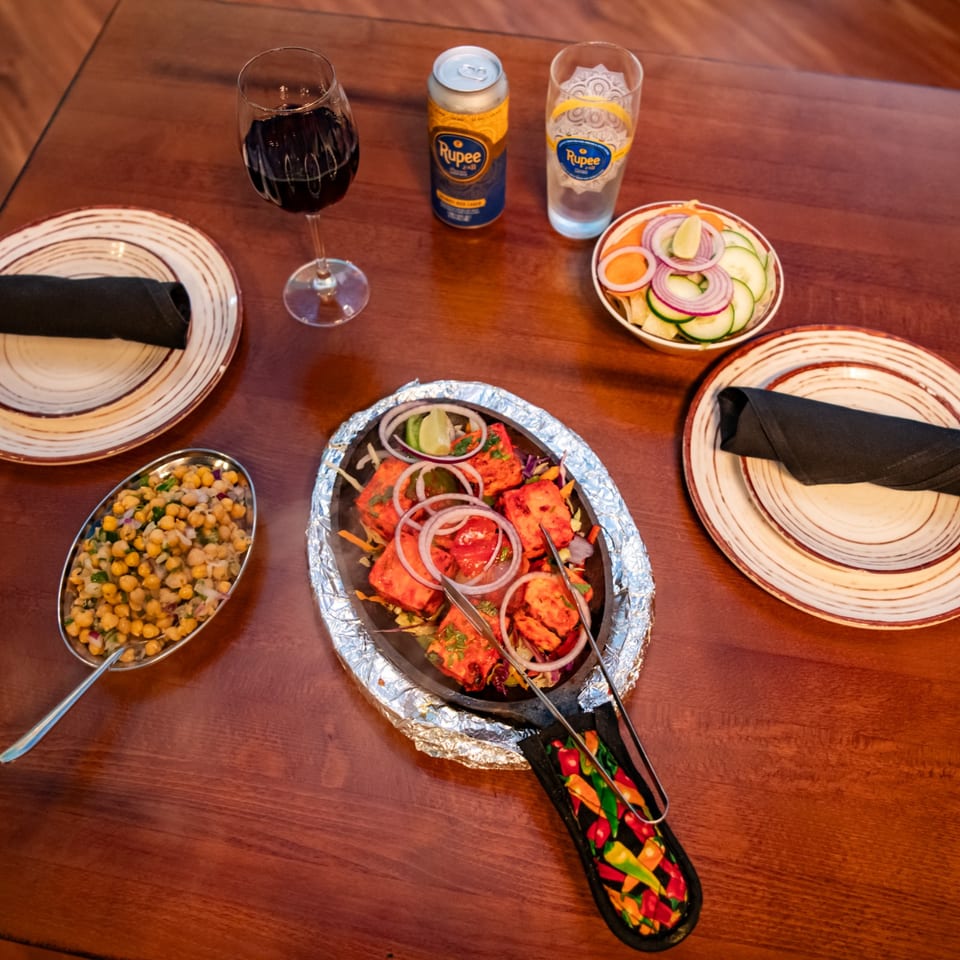 Indian meal spread with grilled paneer, chickpea salad, sliced vegetables, wine, beer, and branded water on a wooden table.