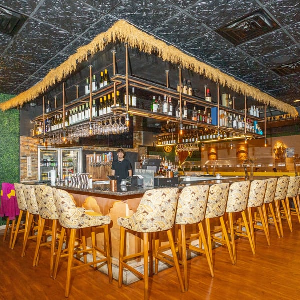 A bartender stands behind a well-stocked bar with a thatched roof and patterned bar stools at Manzil Fine Indian Dining.